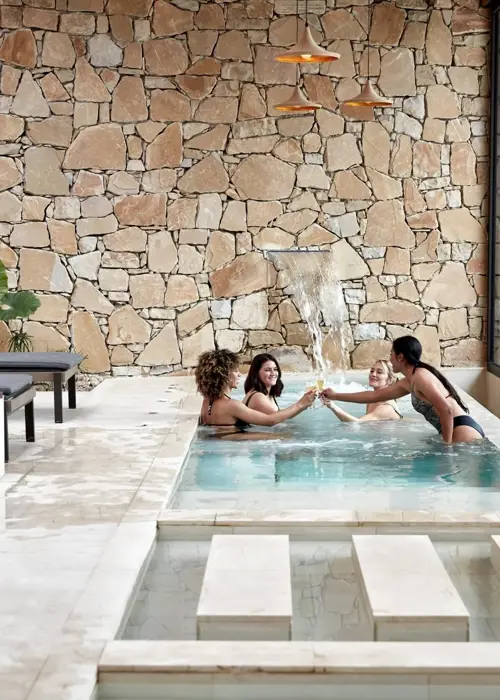 Wide shot of 4 women in thermal pool with champagne glasses at luxury wellness spa bridal package
